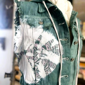 Könnte beinhalten: Eine tealfarbene Jeansweste mit einem weißen und rosa Blumenmuster auf der Vorderseite. Die Weste hat eine Kapuze mit Kordelzug, ausgefranste Kanten und Metallknöpfe. Ein weißer Schal ist im Inneren der Weste zu sehen.