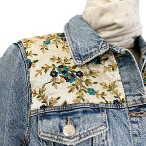 Puede incluir: Chaqueta vaquera azul claro con bordados florales en los hombros. El bordado presenta flores turquesas y doradas sobre un fondo blanquecino. Una bufanda color crema está drapeada sobre el cuello.