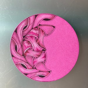 May include: A pink round plate with a black and white intricate design on one side. The design is a series of curved lines and shapes that create a pattern.