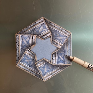 May include: A detailed, geometric art piece in a hexagonal shape, featuring a central star cutout. The design incorporates blue, gray, and gold tones with intricate patterns. A pen is positioned near the artwork, suggesting the creative process.