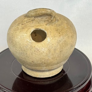 以下が含まれることがあります： 側面に穴が1つ開いた、小さく丸い、オフホワイトの陶器の壺。壺は濃い茶色の木製台座の上に置かれている。