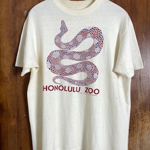 Op de afbeelding: Crèmekleurig T-shirt met een opgerold slangenontwerp in rode, blauwe en witte geometrische patronen. De tekst "HONOLULU ZOO" staat onder de slang, met "OLD ISLAND SHIRTWORKS" eronder.