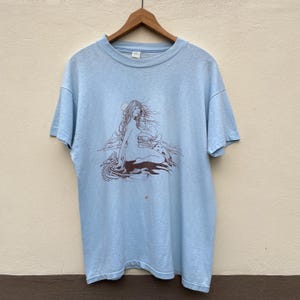 Puede incluir: Camiseta azul claro con una ilustración marrón de una mujer con pelo largo, sentada cerca de una luna y nubes. El gráfico está centrado en la parte delantera de la camiseta, colgada de una percha de madera.