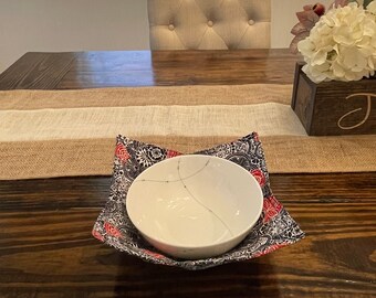 May include: A white ceramic bowl with a black and red floral design on the outside. The bowl is sitting on a dark brown wooden table with a beige runner.