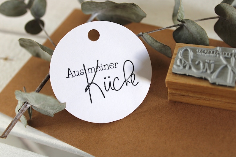Puede incluir: Pegatina redonda blanca con texto negro que dice "Aus meiner K&uuml;che", que se traduce como "De mi cocina" en espa&ntilde;ol. La pegatina tiene un agujero perforado en la parte superior.