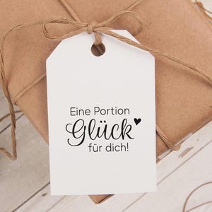 Könnte beinhalten: Ein weißes Geschenkanhänger mit einer schwarzen Schnur darum gebunden. Der Anhänger hat den Text "Eine Portion Glück für dich!" mit einem kleinen schwarzen Herz.