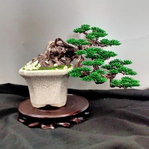 Puede incluir: Una escultura de árbol bonsái de alambre con un tronco de metal retorcido color bronce y follaje de alambre verde vibrante. El árbol está plantado en una maceta de cerámica blanca con acabado craquelado, llena de piedras blancas y musgo verde.