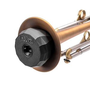 May include: A brass trumpet featuring a matte finish and a black, faceted, geometric mute inserted into the bell. The mute displays the embossed text KGU on its front surface. The instrument includes metallic tubing and circular finger rings.