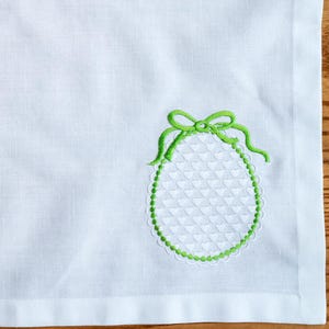 May include: White linen napkin with a green embroidered egg design in the corner. The egg has a textured pattern and is outlined with a green border and bow. The napkin is square and has a finished edge.