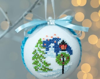 May include: A white Christmas ornament with a cross-stitch design of a winter scene, including a green tree, a house, and a street lamp. The ornament has a blue ribbon and is hanging from a white branch. The background has blurred yellow lights.