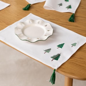 Puede incluir: Mantel individual blanco con árboles de Navidad verdes bordados y una borla verde. Un plato blanco con borde festoneado y recortes de árboles se encuentra sobre el mantel individual. También se ven servilletas a juego.