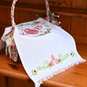 May include: A white linen Easter basket liner with embroidered floral and egg designs. The liner has a fringed edge and a floral wreath with the letter "B" and a cross. The basket handle is wrapped in floral fabric.
