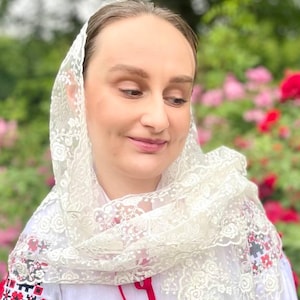 May include: A white embroidered blouse with red and black floral patterns on the sleeves and neckline. A white lace scarf with fringe is draped over the shoulders. The woman is standing in front of a rose bush.