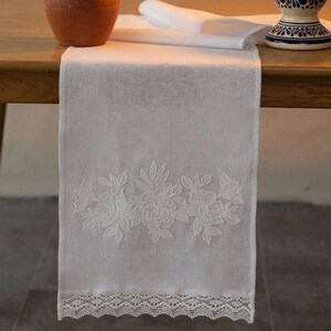 May include: A white linen table runner featuring delicate white floral embroidery and a scalloped lace edge. Beside it sits a terracotta pitcher filled with dried lavender and a ceramic candle holder with a lit white pillar candle.