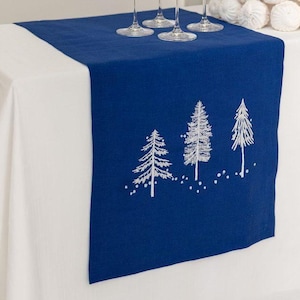 Puede incluir: Un camino de mesa azul marino con un diseño de bosque bordado en blanco. Cuatro copas de vino están dispuestas sobre el camino de mesa, que se coloca sobre un mantel blanco. Un árbol decorativo de postre está en el fondo.