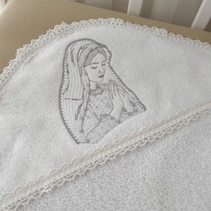 Może przedstawiać: Biały ręcznik z kapturem, ozdobiony koronką i haftem przedstawiającym Matkę Boską z dłońmi złożonymi do modlitwy.