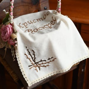 Puede incluir: Una cubierta de cesta de Pascua de lino blanco con texto bordado y ramas de sauce. La cubierta tiene un delicado ribete de encaje y está decorada con flores rosas y vegetación en el asa.