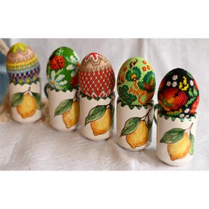 Puede incluir: Seis huevos de Pascua decorativos con intrincados abalorios en varios patrones y colores, descansan sobre pequeñas tazas de cerámica. Las tazas presentan un diseño de limón pintado a mano, añadiendo un toque rústico. Los huevos muestran una variedad de diseños.