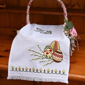Può includere: Fodera per cestino pasquale bianca con uova di Pasqua ricamate e rami di salice. Le uova sono decorate con motivi floreali rossi e gialli. Il manico del cestino è avvolto in tessuto floreale.