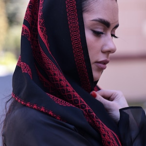 May include: A black headscarf featuring intricate red geometric embroidery along the borders and across the fabric. The scarf is draped over the head, showcasing a detailed pattern of repeating lines and shapes in a vibrant red thread on black.