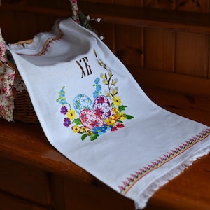 May include: White embroidered Easter towel with colourful floral and egg designs. The towel has a decorative border and the letters "ХВ" embroidered in brown. The towel is draped over a wooden surface.