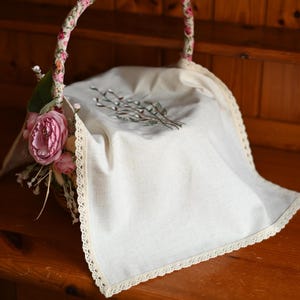 Puede incluir: Una cesta decorativa con un asa floral y un paño de lino color crema. El paño está bordado con una ramita de sauce y adornado con encaje. Rosas rosas y vegetación adornan el lateral de la cesta.