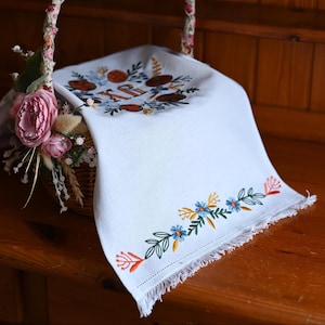 May include: A white linen towel with embroidered floral designs, including blue, orange, and yellow flowers, and green leaves. The towel is draped over a wicker basket decorated with flowers. The towel has a fringed edge.