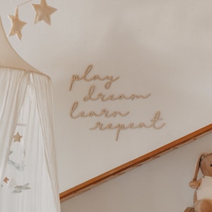 Puede incluir: Una cama con dosel blanco con una guirnalda en forma de estrella colgando encima. La pared detr&aacute;s de la cama tiene un letrero de madera que dice "play dream learn repeat". Hay un conejito de peluche y un arco&iacute;ris de madera en un estante debajo del letrero.