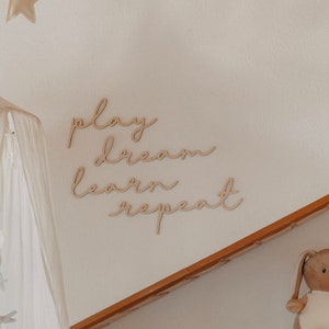 Puede incluir: Decoraci&oacute;n de pared de madera con las palabras "play dream learn repeat" en una fuente cursiva. La decoraci&oacute;n est&aacute; montada en una pared blanca con un dosel blanco colgando en el fondo.