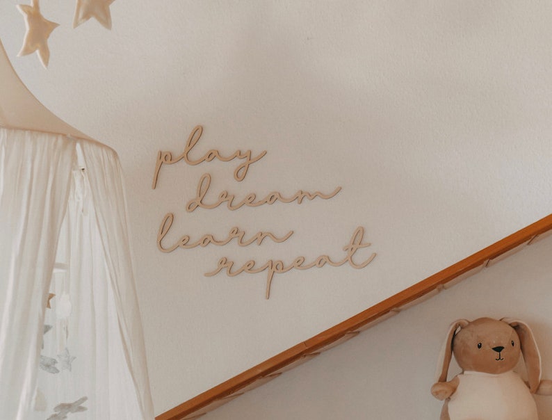 Puede incluir: Decoraci&oacute;n de pared de madera con las palabras "play dream learn repeat" en una fuente cursiva. La decoraci&oacute;n est&aacute; montada en una pared blanca con un dosel blanco colgando en el fondo.