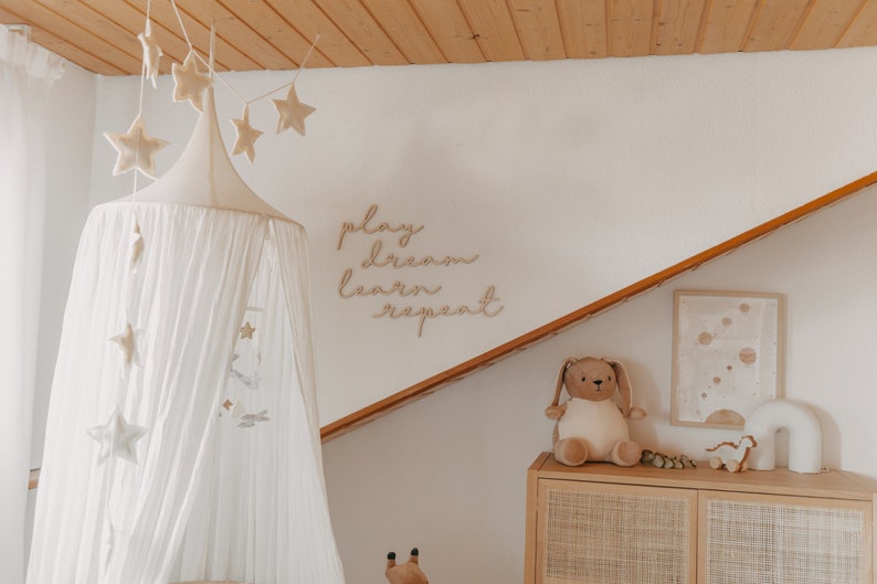 Puede incluir: Una cama con dosel blanco con una guirnalda en forma de estrella colgando encima. La pared detr&aacute;s de la cama tiene un letrero de madera que dice "play dream learn repeat". Hay un conejito de peluche y un arco&iacute;ris de madera en un estante debajo del letrero.