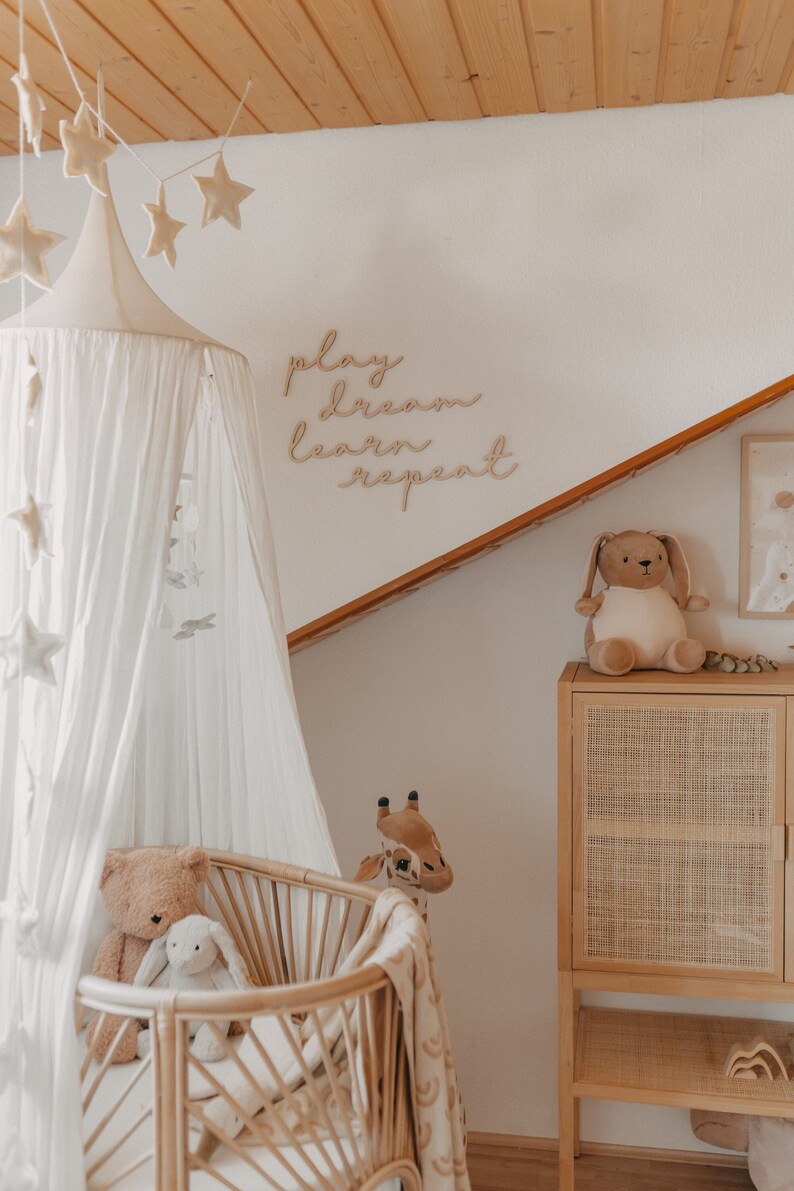 Puede incluir: Una cuna con dosel blanco con una cuna de mimbre, un oso de peluche marr&oacute;n, un conejito blanco y una jirafa de peluche. La pared detr&aacute;s de la cuna tiene un letrero de madera que dice "play dream learn repeat".