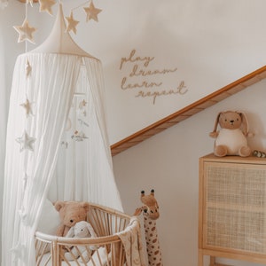 Puede incluir: Una cuna con dosel blanco con una jirafa de peluche y un oso de peluche. La cuna est&aacute; decorada con un letrero de madera que dice "play dream learn repeat".