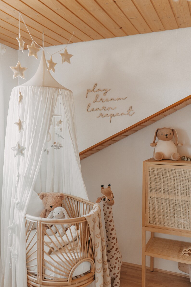 Puede incluir: Una cuna con dosel blanco con una jirafa de peluche y un oso de peluche. La cuna est&aacute; decorada con un letrero de madera que dice "play dream learn repeat".