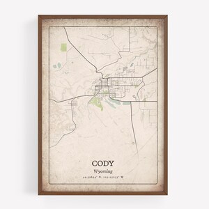 May include: Framed map print of Cody, Wyoming, featuring a detailed street layout and surrounding areas. The map has a vintage aesthetic with a beige background and brown frame. The city name and coordinates are displayed at the bottom.