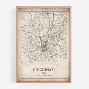 Puede incluir: Una impresión enmarcada del mapa de Cincinnati, Ohio, con una red de carreteras detallada en líneas negras sobre un fondo beige. El nombre de la ciudad y las coordenadas están impresos en la parte inferior. El marco es de color marrón claro.
