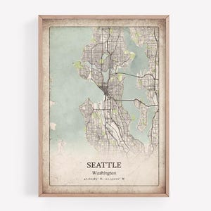 May include: A framed map print of Seattle, Washington, with a vintage aesthetic. The map features a detailed street layout in grey and black, set against a pale green and beige background. The city name and coordinates are printed at the bottom.