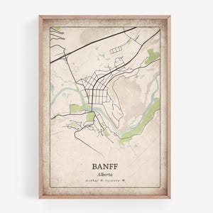 May include: A framed map print of Banff, Alberta, featuring a detailed street layout and surrounding geographical features. The map is in black and white with green accents, set against a vintage-style beige background. The town's name and coordinates are printed at the bottom.
