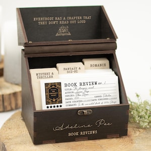 Op de afbeelding: Een donkerbruine houten boekrecensiebox met een scharnierend deksel, met boekrecensiekaarten. De doos is gegraveerd met tekst, waaronder "EVERYBODY HAS A CHAPTER THAT THEY DON'T READ OUT LOUD" en "Adeline Poe BOOK REVIEWS". De doos staat op een houten schijf.