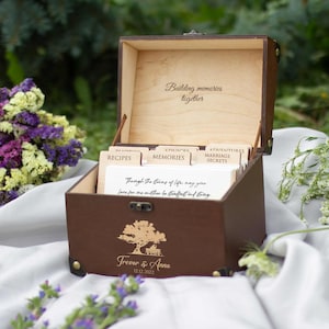 May include: A brown wooden memory box, open to show labeled dividers for recipes, memories, advice, and marriage secrets. The box has a tree design and the names Trevor & Anna. The text "Building memories together" is inside the lid. The box is on a white cloth.