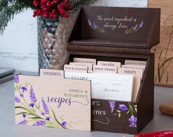 Peut inclure: Une boîte à recettes en bois avec un motif floral de lavande. La boîte a une section supérieure avec le texte "L'ingrédient secret est toujours l'amour." et une section inférieure avec des porte-cartes de recettes étiquetés "Desserts", "Pâtes et céréales", "Poisson et fruits de mer", "Salades", "Plats principaux", "Pain et pâtisserie", et "Viandes". La boîte à recettes est étiquetée "Recettes de Joshua et Elizabeth".