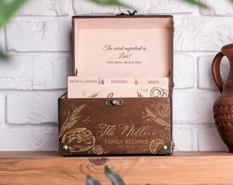 May include: A wooden recipe box with the text "The Millers Family Recipes EST 2025" and "The secret ingredient is Love!" The box is open, revealing recipe dividers for pasta, desserts, and meats. A ceramic jug sits to the right.