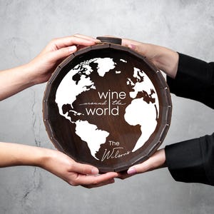 May include: A dark wooden barrel with a white world map design and the text "wine around the world". The barrel is held by two people. The text "The Wilsons" is also on the barrel.