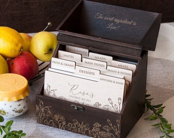 May include: A wooden recipe box with a dark brown finish and a floral design. The box has a hinged lid and is divided into sections for different types of recipes. The lid has the text "The secret ingredient is Love!" engraved on it. The box is labelled with categories such as "Seafood", "Sauces", "Pasta & Grains", "Salads", "Main Dishes", "Desserts", "Meats", "Breads & Pastries", and "Appetizers & Soups".