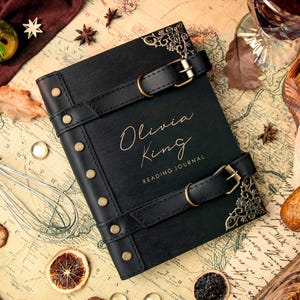 Op de afbeelding: Een zwart leren leesdagboek met de naam "Olivia King" in gouden letters. Het dagboek heeft twee zwarte leren riemen met gouden gespen en decoratieve hoekaccenten. Het dagboek ligt op een kaart met andere items.