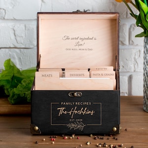 May include: A black wooden recipe box with a hinged lid, featuring the text "The Hochkins" and "Family Recipes". Inside, labeled dividers organise recipes. The box is decorated with brass-coloured corner accents and a floral design. The lid has the text "The secret ingredient is Love!"