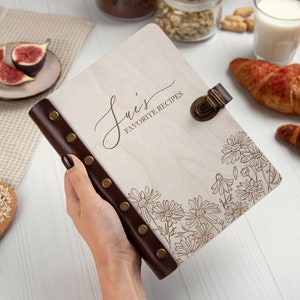 May include: A white wooden recipe book with a brown leather spine and metal clasps. The book has a floral design and the text "Sue's Favorite Recipes" on the front cover.