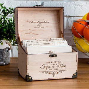 May include: Wooden recipe box with an open lid, featuring the text "The secret ingredient is Love" and "Grandma Lilian." The box is personalized with "Sofia & Max" and "June 7, 2021." It contains labeled dividers for recipes.