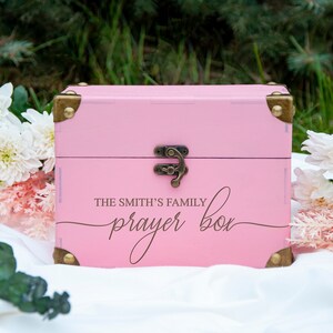 May include: A pink wooden box with a brass latch and hinges. The box has the text "The Smith's Family Prayer Box" written on the front in a cursive font.