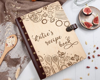 May include: A wooden recipe book with a brown leather spine and a clasp. The cover is engraved with a design of kitchen utensils and ingredients, and the text "Katie's recipe book" is written in cursive.